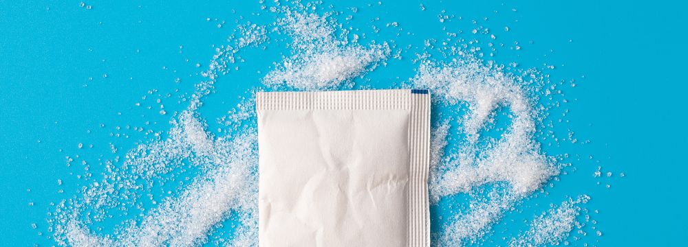 Packet of sweetener sprinkled on blue background illustrates concept of sugar versus artificial sweetener for health