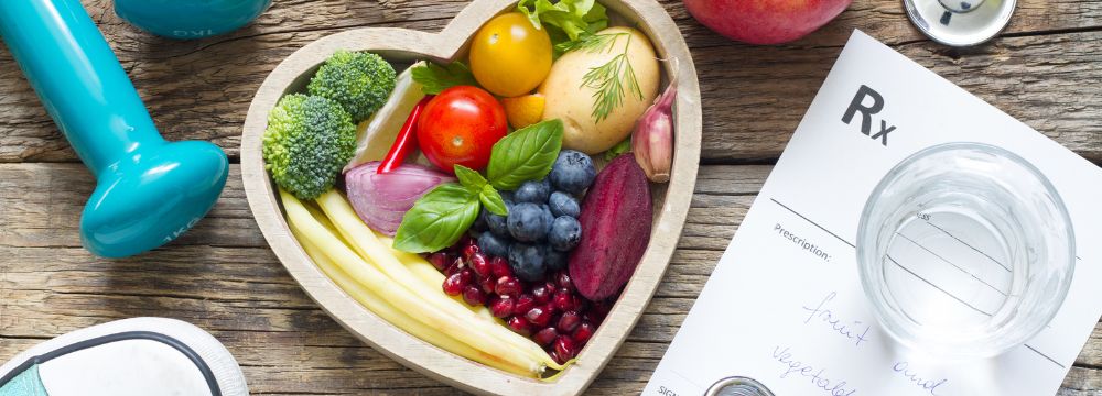 Heart shaped bowl filled with fruit and veggies next to prescription paper and blue dumbell