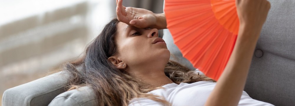 Woman lying on couch sweating with tand on forehead holding paper fan 